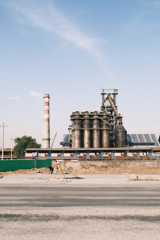 An industrial facility with large cylindrical metal structures and a tall chimney that has red and white stripes. The foreground shows a person in an orange vest working with a tripod and surveying equipment. The sky is clear with a few clouds, and the area around the facility appears under construction or renovation.