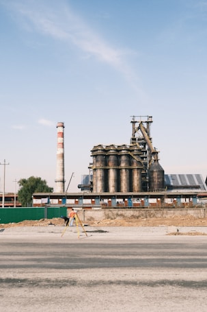 An industrial facility with large cylindrical metal structures and a tall chimney that has red and white stripes. The foreground shows a person in an orange vest working with a tripod and surveying equipment. The sky is clear with a few clouds, and the area around the facility appears under construction or renovation.