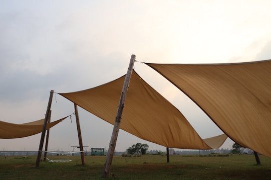 Large tan canvases are stretched between wooden poles on a grassy field. The sky is overcast with a soft, diffuse light. In the background, trees are visible and a structure that resembles a trailer or bus.