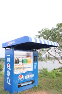 A blue Pepsi vending machine is situated outdoors on a grassy area. It has a striped blue and white awning above it and displays various drinks in clear compartments. Behind the vending machine, trees and a body of water are visible under a cloudy sky.