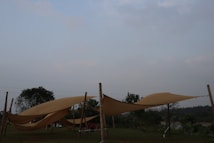 Several beige fabric canopies are stretched between wooden poles in an outdoor setting. The scene includes grassy areas and scattered trees with a pale sky in the background.