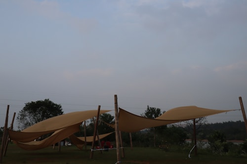 Several beige fabric canopies are stretched between wooden poles in an outdoor setting. The scene includes grassy areas and scattered trees with a pale sky in the background.
