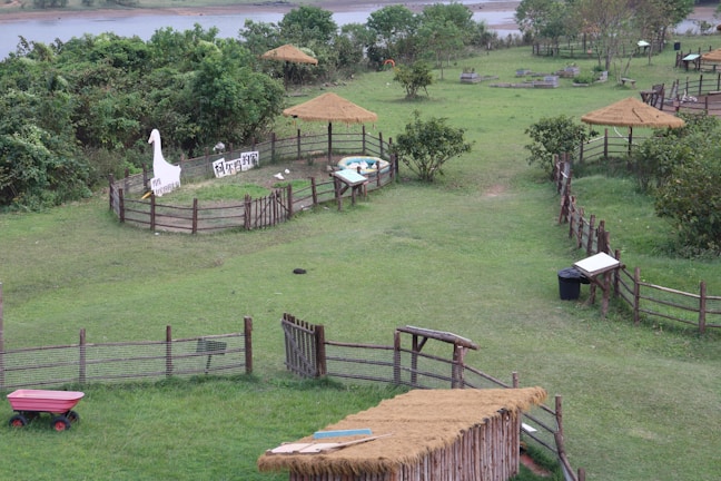 A serene outdoor setting with a grassy field enclosed by wooden fences. There are several thatch-roofed structures and a penned area with a large cutout of a duck. Trees and shrubs are scattered throughout the landscape, and a river is visible in the distance. A red wagon is positioned on the left side and there are some signs with text in an Asian script.