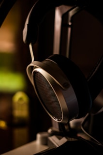 A close-up of a pair of over-ear headphones with large padded ear cups and a metallic frame. The focus is on the mesh grill of one ear cup, with a blurred background featuring warm lighting.