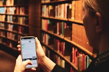 A person is holding a smartphone displaying a digital library application while surrounded by shelves filled with books. The shelves are arranged in rows, and the lighting creates a warm and cozy atmosphere. The individual appears to be browsing library services or book information on their phone.