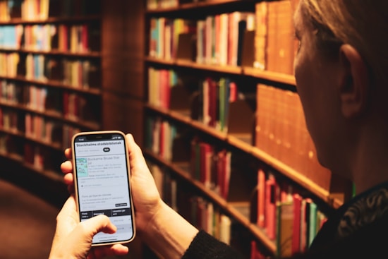 A person is holding a smartphone displaying a digital library application while surrounded by shelves filled with books. The shelves are arranged in rows, and the lighting creates a warm and cozy atmosphere. The individual appears to be browsing library services or book information on their phone.