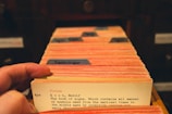 A hand flips through a collection of vintage library catalog cards, each with typed text and some colored tabs. The focus is on one card that has text describing a book titled 'The Book of Signs.' The cards are housed in a wooden drawer setup, suggesting a traditional library or archival setting.