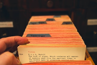 A hand flips through a collection of vintage library catalog cards, each with typed text and some colored tabs. The focus is on one card that has text describing a book titled 'The Book of Signs.' The cards are housed in a wooden drawer setup, suggesting a traditional library or archival setting.