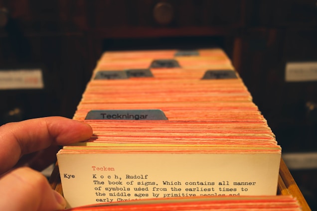 A hand flips through a collection of vintage library catalog cards, each with typed text and some colored tabs. The focus is on one card that has text describing a book titled 'The Book of Signs.' The cards are housed in a wooden drawer setup, suggesting a traditional library or archival setting.