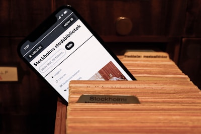 A smartphone screen displays a webpage for the Stockholm City Library, featuring a search bar and library-related options. The phone is placed inside a wooden card catalog drawer filled with organized index cards.