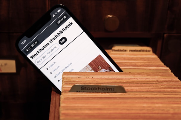 A smartphone screen displays a webpage for the Stockholm City Library, featuring a search bar and library-related options. The phone is placed inside a wooden card catalog drawer filled with organized index cards.