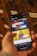 A person holding a smartphone displaying a webpage with a Scandinavian library streaming service. The screen shows two video thumbnails, one with a TV displaying a program and the other with a woman holding a book. The background features a wooden drawer set with labels.