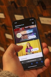 A person holding a smartphone displaying a webpage with a Scandinavian library streaming service. The screen shows two video thumbnails, one with a TV displaying a program and the other with a woman holding a book. The background features a wooden drawer set with labels.
