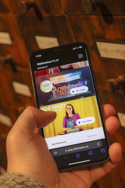 A person holding a smartphone displaying a webpage with a Scandinavian library streaming service. The screen shows two video thumbnails, one with a TV displaying a program and the other with a woman holding a book. The background features a wooden drawer set with labels.