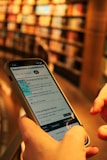A person is holding a smartphone in their hands, navigating an online library or e-book application with blurred bookshelves in the background, giving a sense of depth and focus on the technology.