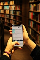 A person is holding a smartphone in both hands, accessing a library's online catalog app, with shelves filled with books in a dimly lit library setting. The phone screen displays text and images related to Stockholm's public library.