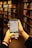 A person is holding a smartphone in both hands, accessing a library's online catalog app, with shelves filled with books in a dimly lit library setting. The phone screen displays text and images related to Stockholm's public library.
