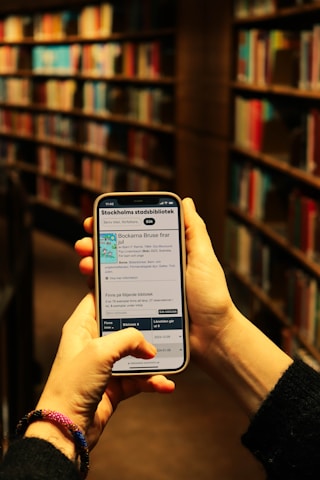 A person is holding a smartphone in both hands, accessing a library's online catalog app, with shelves filled with books in a dimly lit library setting. The phone screen displays text and images related to Stockholm's public library.