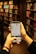 A person is holding a smartphone in both hands, accessing a library's online catalog app, with shelves filled with books in a dimly lit library setting. The phone screen displays text and images related to Stockholm's public library.