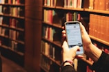 A person is holding a smartphone in a library setting. The phone screen displays information, likely from a library app or website, and bookshelves filled with books are visible in the background.