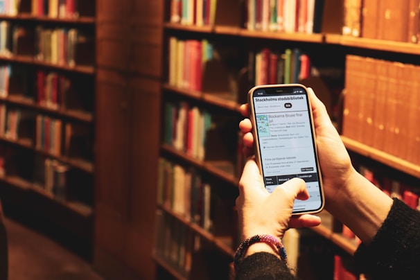 A person is holding a smartphone in a library setting. The phone screen displays information, likely from a library app or website, and bookshelves filled with books are visible in the background.