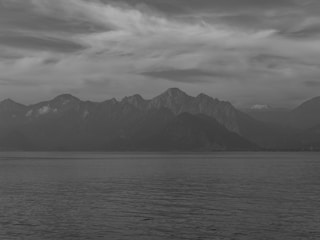 A monochrome image of a serene landscape features a calm body of water in the foreground with distant mountain ranges under a sky filled with wispy clouds. The absence of color emphasizes the contrast between the dark water and the lighter sky.