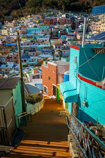 A vibrant neighborhood with a multitude of colorful houses built closely together on a hillside. The houses are painted in bright shades of blue, green, yellow, pink, and orange. There are narrow, winding stairways and pathways leading through the area, lined with metal railings. Electrical wires crisscross above the rooftops, indicating a densely populated urban neighborhood.