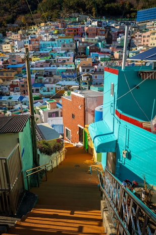 A vibrant neighborhood with a multitude of colorful houses built closely together on a hillside. The houses are painted in bright shades of blue, green, yellow, pink, and orange. There are narrow, winding stairways and pathways leading through the area, lined with metal railings. Electrical wires crisscross above the rooftops, indicating a densely populated urban neighborhood.