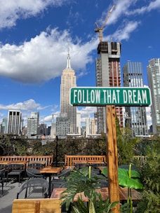 A rooftop scene featuring a 'Follow That Dream' sign in the foreground, surrounded by lush green plants and outdoor seating. The skyline showcases modern skyscrapers and the Empire State Building under a bright blue sky with scattered clouds. A construction crane is visible on one of the buildings.