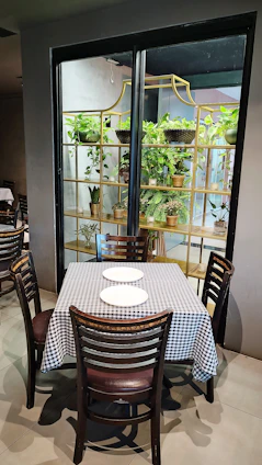A small dining table with a black and white checkered tablecloth is set with two white plates. The table is surrounded by four wooden chairs with dark cushions. In the background, there is a large shelf filled with various potted green plants, adding a touch of nature to the otherwise minimalistic room.