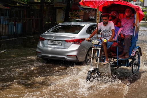 A man is pedaling a rickshaw carrying two passengers through a flooded street. The passengers are sheltered under a red umbrella, and there is a silver car driving through the same floodwaters next to the rickshaw. In the background, there is a metal fence and some buildings.