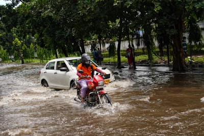 A flooded street with a person wearing a helmet riding a red motorcycle through the water, while a white car is partially submerged. Trees line the road, and a few pedestrians appear to be walking cautiously along the sidewalk.
