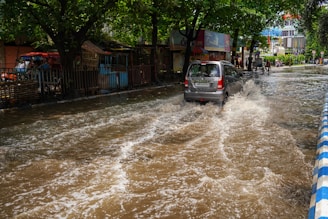 A car drives through a flooded street with water splashing around in an urban area. Trees line the side of the street, and there are various structures and signs in the background.
