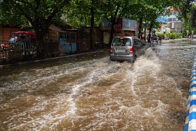 A car drives through a flooded street with water splashing around in an urban area. Trees line the side of the street, and there are various structures and signs in the background.