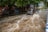 A car drives through a flooded street with water splashing around in an urban area. Trees line the side of the street, and there are various structures and signs in the background.