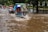 A flooded street with several vehicles, including motorbikes and auto rickshaws, navigating through the water. People are seen riding the vehicles, wearing helmets, and the surrounding area is lush with green trees.