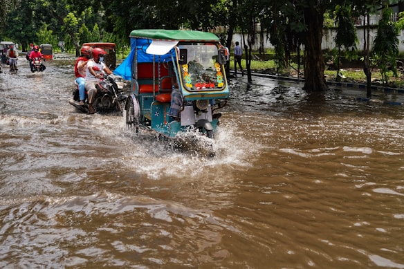 A flooded street with several vehicles, including motorbikes and auto rickshaws, navigating through the water. People are seen riding the vehicles, wearing helmets, and the surrounding area is lush with green trees.