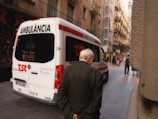 An ambulance is parked on a narrow urban street with an elderly man walking past it. The street is flanked by tall, older buildings with balconies, and a few people can be seen walking or on bicycles further down the street.