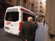 An ambulance is parked on a narrow urban street with an elderly man walking past it. The street is flanked by tall, older buildings with balconies, and a few people can be seen walking or on bicycles further down the street.