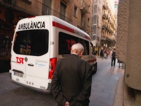 An ambulance is parked on a narrow urban street with an elderly man walking past it. The street is flanked by tall, older buildings with balconies, and a few people can be seen walking or on bicycles further down the street.