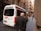 An ambulance is parked on a narrow urban street with an elderly man walking past it. The street is flanked by tall, older buildings with balconies, and a few people can be seen walking or on bicycles further down the street.