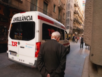 An ambulance is parked on a narrow urban street with an elderly man walking past it. The street is flanked by tall, older buildings with balconies, and a few people can be seen walking or on bicycles further down the street.
