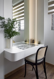 A modern, minimalist vanity setup with a glass-topped white table featuring organized compartments. There is a black chair with a beige cushion. A green plant in a white vase is placed on the left side of the vanity. LED lighting creates a warm glow behind the mirror. Blinds partially cover the windows, allowing natural light into the room. Small decorative items are neatly arranged on the vanity.