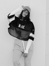 A young woman is posing against a plain wall, wearing a cap, a sporty oversized mesh shirt with long sleeves underneath, and casual sweatpants. She holds the brim of the cap with one hand and her hair is partially covering her face.