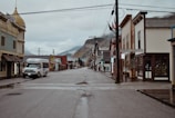 A quiet, empty street in a small town lined with historic buildings showcasing a variety of local businesses. A shuttle bus is parked on the left side of the street, which is wet from recent rain. The overcast sky and surrounding mountains contribute to the serene atmosphere.
