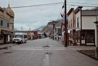 A quiet, empty street in a small town lined with historic buildings showcasing a variety of local businesses. A shuttle bus is parked on the left side of the street, which is wet from recent rain. The overcast sky and surrounding mountains contribute to the serene atmosphere.