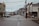 A quiet, empty street in a small town lined with historic buildings showcasing a variety of local businesses. A shuttle bus is parked on the left side of the street, which is wet from recent rain. The overcast sky and surrounding mountains contribute to the serene atmosphere.