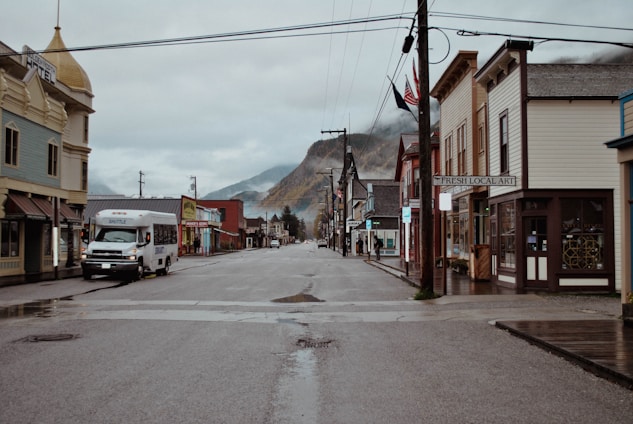 A quiet, empty street in a small town lined with historic buildings showcasing a variety of local businesses. A shuttle bus is parked on the left side of the street, which is wet from recent rain. The overcast sky and surrounding mountains contribute to the serene atmosphere.