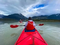 Brightly colored kayaks glide across a serene lake surrounded by majestic mountains under a cloudy sky. Two people are paddling, wearing red life jackets and casually dressed for a cool day. The water is calm with gentle ripples, creating a peaceful scene in a natural landscape.