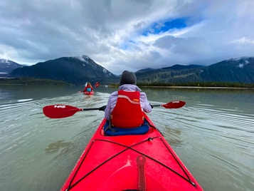 Brightly colored kayaks glide across a serene lake surrounded by majestic mountains under a cloudy sky. Two people are paddling, wearing red life jackets and casually dressed for a cool day. The water is calm with gentle ripples, creating a peaceful scene in a natural landscape.